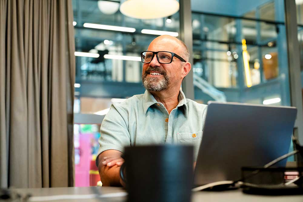Leende äldre affärsman med glasögon och pipskägg deltar i onlinemöte vid laptop på modernt kontor